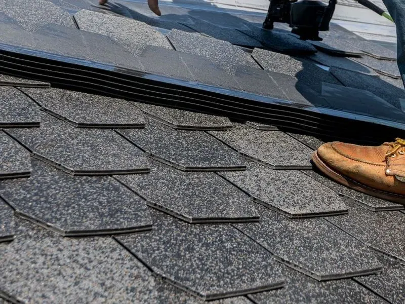 Close-up of a worker's boot and nail gun installing asphalt shingles on a roof